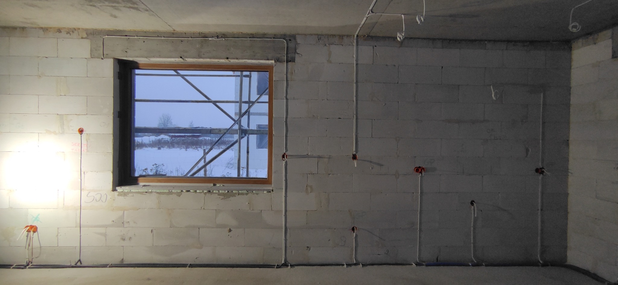 Surowy pokój z białego bloczka, widoczne przewody elektryczne w peszlach prowadzone po ścianach, okno z widokiem na zimowy krajobraz i rusztowanie.