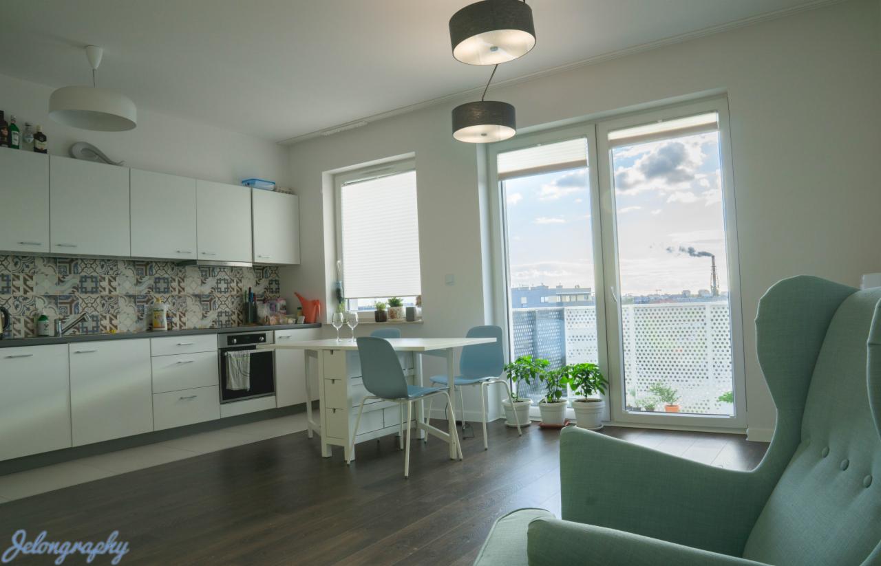 Jasne wnętrze kuchni z białymi szafkami, wzorzystą mozaiką nad blatem, stołem jadalnym z krzesłami, dużymi oknami z widokiem na miasto i komin fabryczny w oddali oraz fragment turkusowego fotela...