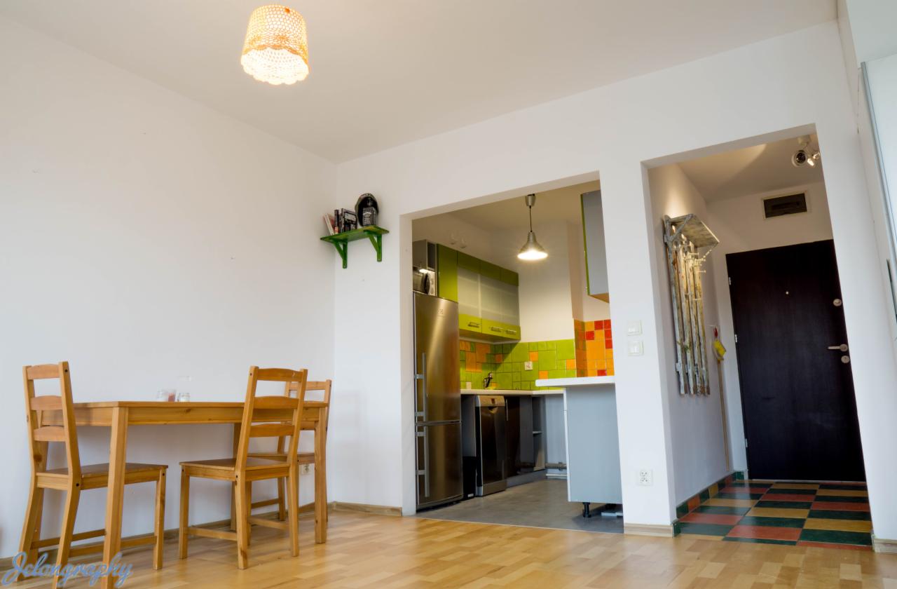 Jasne, przestronne wnętrze mieszkania z widokiem na jadalnię z drewnianym stołem i krzesłami, otwartą kuchnię z zielonymi szafkami i kolorowymi kafelkami oraz przedpokój z ciemnymi drzwiami...