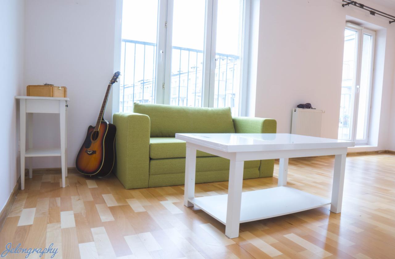 Jasny pokój dzienny z zieloną sofą, białym stolikiem kawowym, drewnianą podłogą i gitarą opartą o ścianę obok okna.
