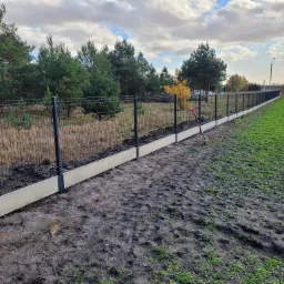 Długi odcinek panelowego ogrodzenia z podmurówką, zamontowany na grząskim terenie, z widoczną czerwoną łopatą opartą o ogrodzenie oraz białym busem w oddali.