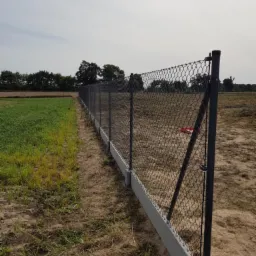 Długa siatka ogrodzeniowa z betonową podmurówką, widoczna na tle pola w Bydgoszczy. Słupki metalowe, siatka w ciemnym kolorze.