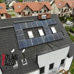 Panele słoneczne umieszczone na dachu domu z dachówką w kolorze antracytowym, widoczne okna dachowe i kominy, w tle zabudowa szeregowa i zadbane ogrody.