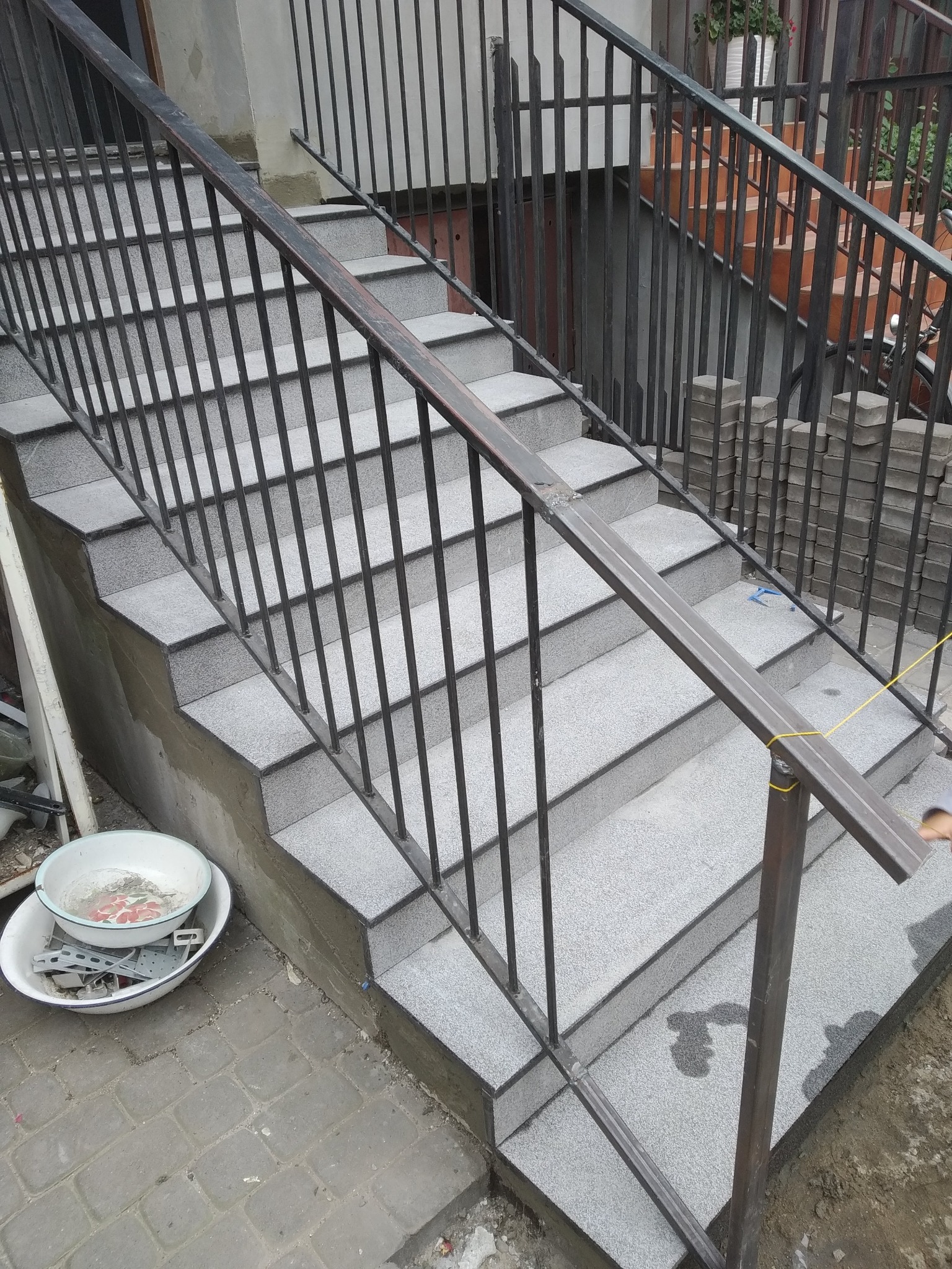 Zewnętrzne schody wyłożone szarymi płytkami imitującymi kamień, z metalową balustradą, w trakcie montażu lub po zakończeniu prac, z widocznymi śladami wody na powierzchni.