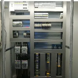 Rozdzielnica elektryczna w metalowej obudowie z widocznymi zabezpieczeniami, licznikami i okablowaniem, zainstalowana w pomieszczeniu.