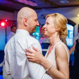 Para młoda, on łysy w białej koszuli, ona blondynka w sukni ślubnej, patrzą na siebie na parkiecie podczas wesela, w tle goście i kolorowe światła.