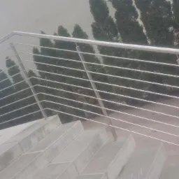 Nowoczesna balustrada ze stali nierdzewnej z poziomymi prętami, zamontowana na betonowych schodach zewnętrznych, z widokiem na zielone tuje w tle.