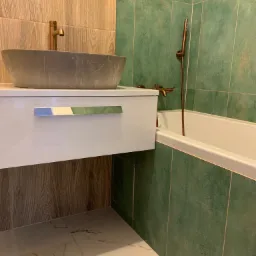 Nowoczesna łazienka z zielonymi płytkami, marmurową umywalką nablatową, złotą armaturą i białymi szafkami z chromowanymi uchwytami.