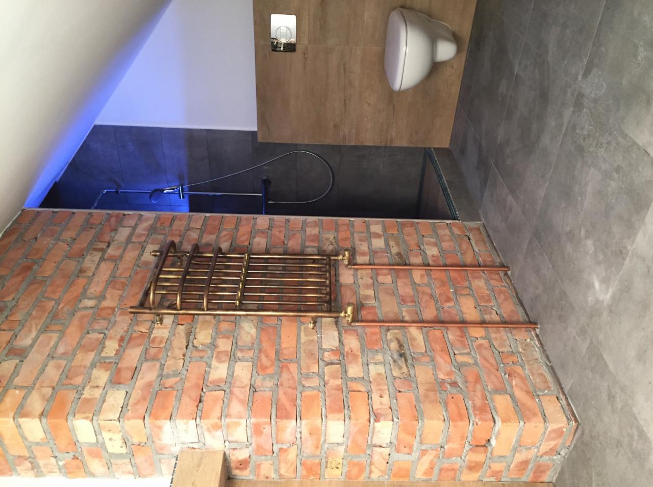 Łazienka na poddaszu z ceglaną ścianą, miedzianym grzejnikiem drabinkowym i instalacją wodną, szare płytki na podłodze i ścianach, biała toaleta pod skosem dachu z niebieskim oświetleniem.