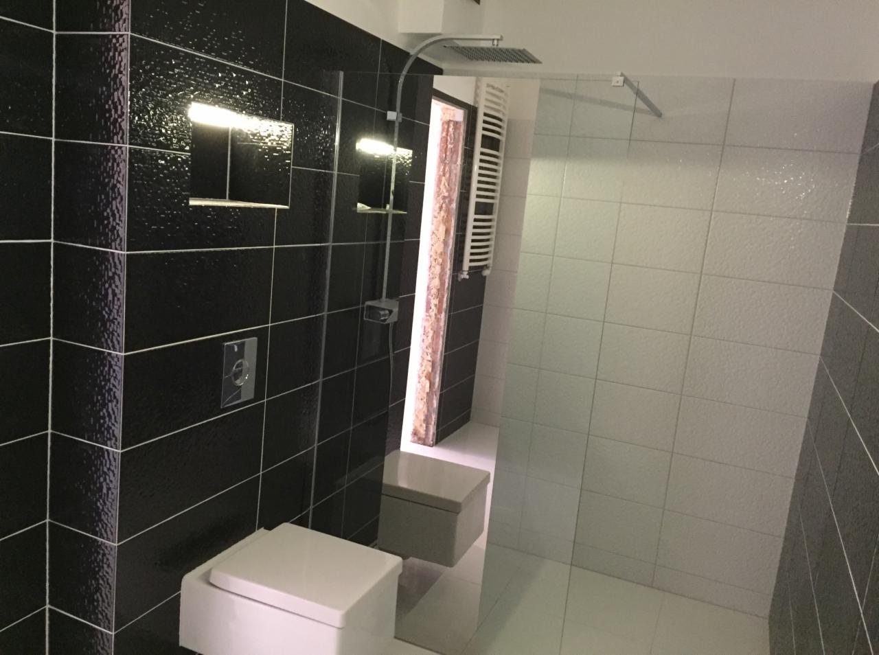 Nowoczesna łazienka z czarno-białymi płytkami, wiszącą toaletą i deszczownicą, oświetlona ciepłym światłem.