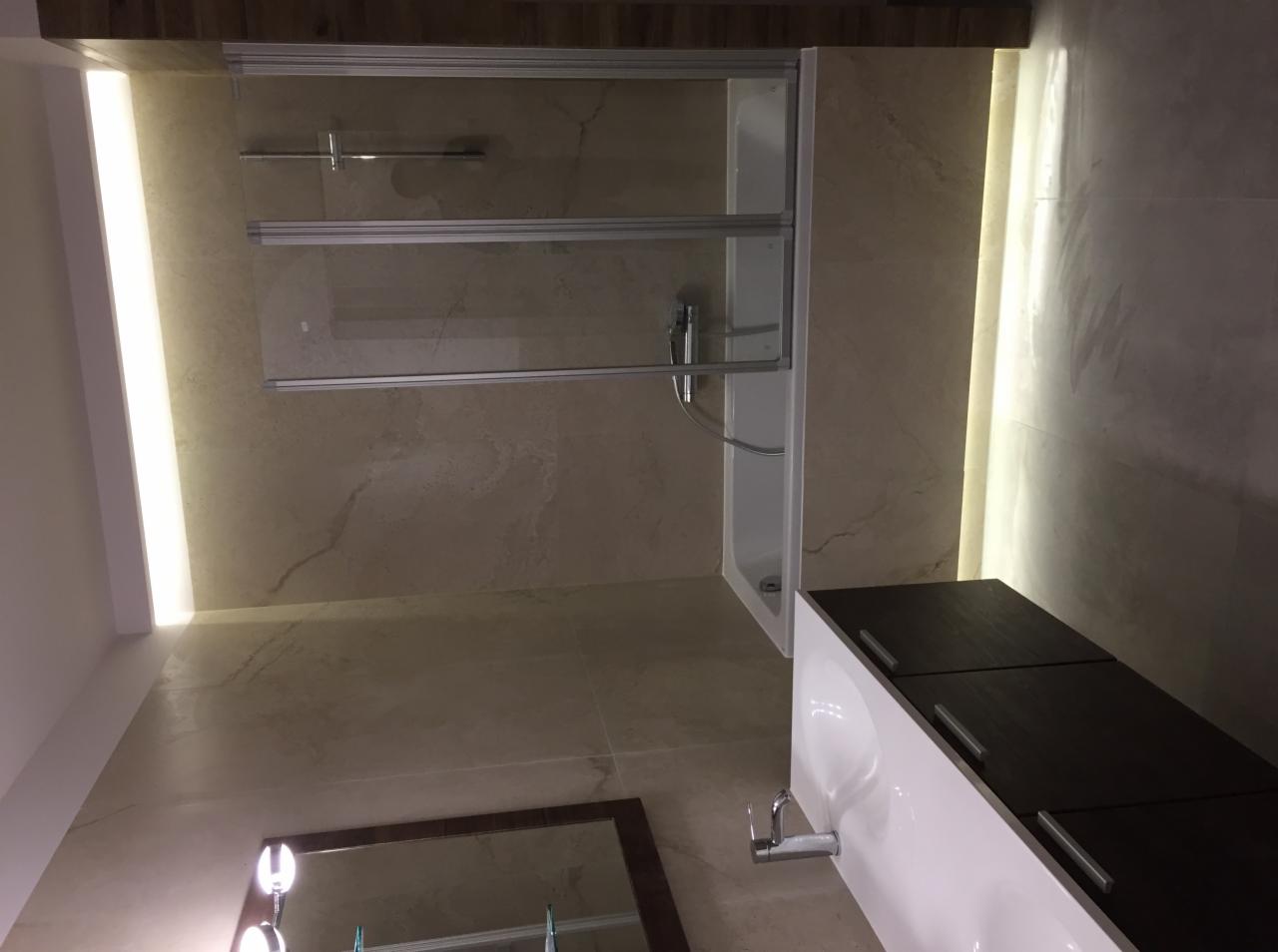 Nowoczesna łazienka z beżowymi płytkami, kabiną prysznicową z przesuwanymi drzwiami, podświetleniem LED i umywalką z szafką w kolorze białym i ciemnego drewna.