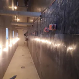 Wnętrze hali w trakcie prac nad posadzką: widoczna wylewka na podłożu, zabezpieczona folią ściana, ekipa w oddali oraz narzędzia pomiarowe.