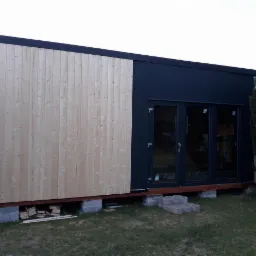 Nowoczesny budynek modułowy z elewacją z surowego drewna i czarnymi panelami, posadowiony na betonowych bloczkach, widoczne ułożone bloczki schodowe.