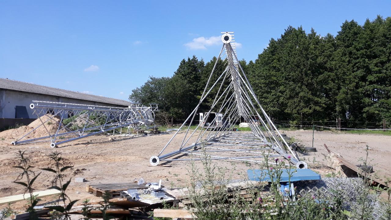 Konstrukcja metalowej wieży telekomunikacyjnej w trakcie montażu na placu budowy; widoczne elementy składowe, podłoże gruntowe i budynek gospodarczy w tle.