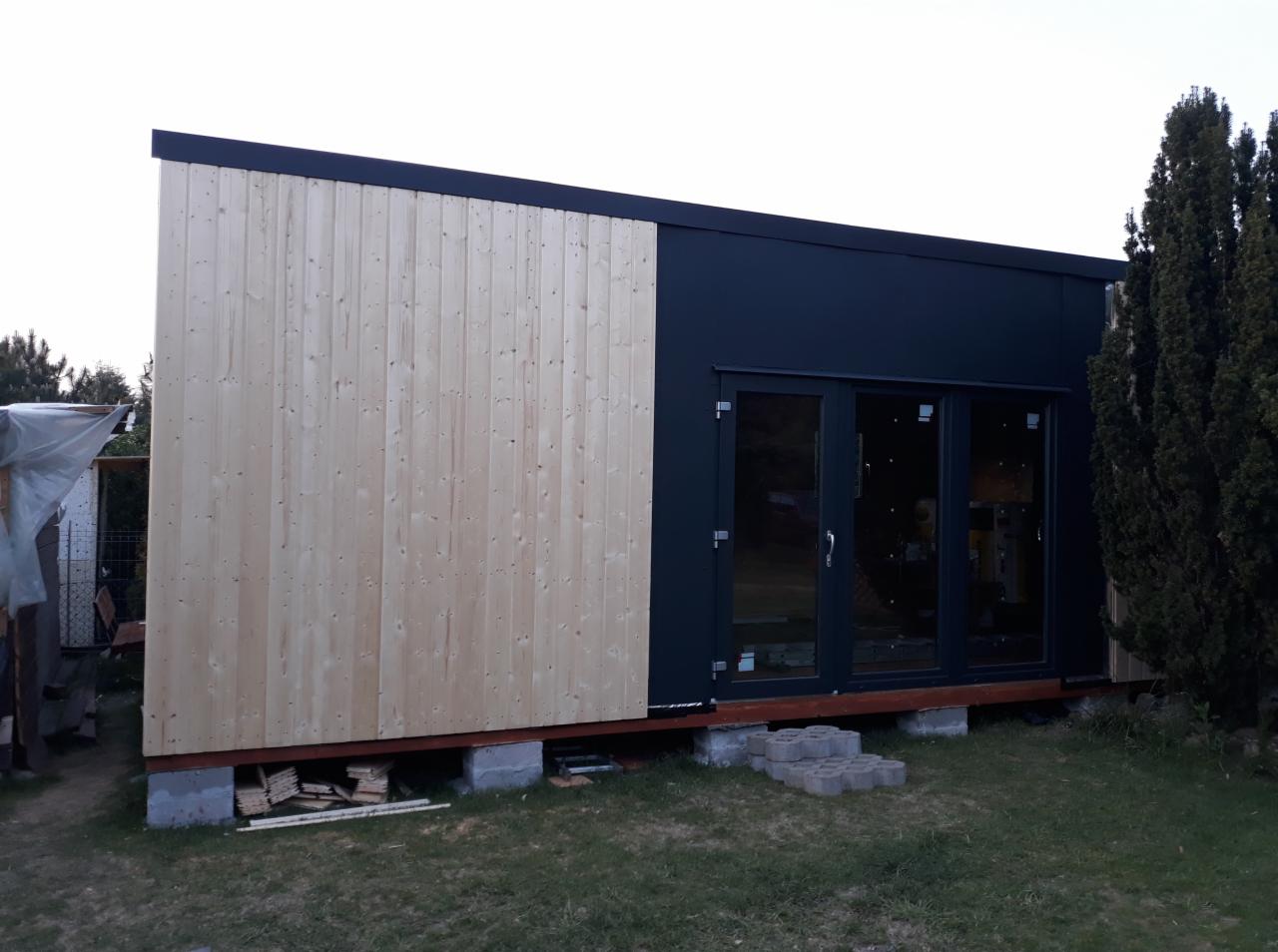 Nowoczesny budynek modułowy z elewacją z surowego drewna i czarnymi panelami, posadowiony na betonowych bloczkach, widoczne ułożone bloczki schodowe.