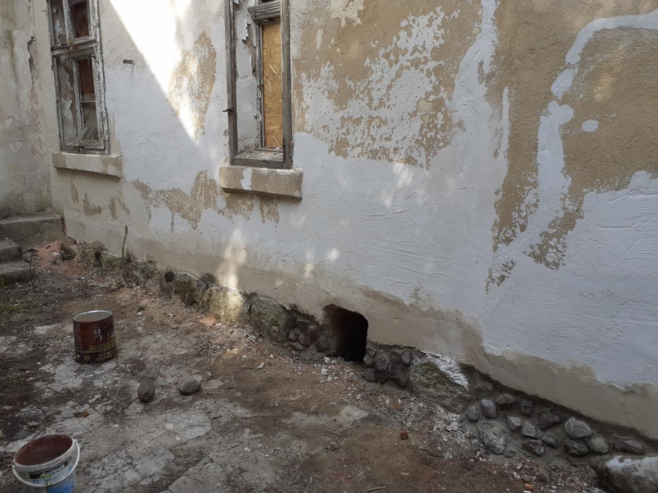 Fragment ściany budynku w trakcie prac: częściowo otynkowana i pomalowana na biało, z widocznymi śladami starego tynku, zaklejonymi oknami i odsłoniętym fundamentem z kamieni.