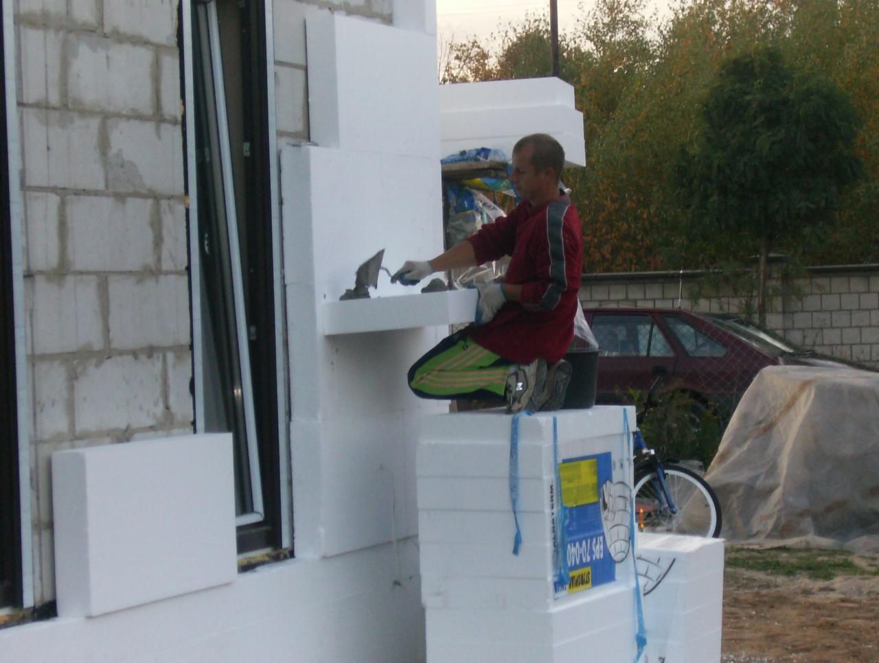 Pracownik w czerwonej bluzie aplikuje zaprawę na styropian podczas ocieplania elewacji budynku, widoczny fragment okna, bloczki oraz stos białego styropianu na pierwszym planie, w tle zaparkowany...