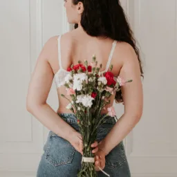 Kobieta odwrócona tyłem, trzymająca za plecami bukiet czerwonych, białych i różowych goździków, ubrana w dżinsy i koronkowy biustonosz na tle białych drzwi.