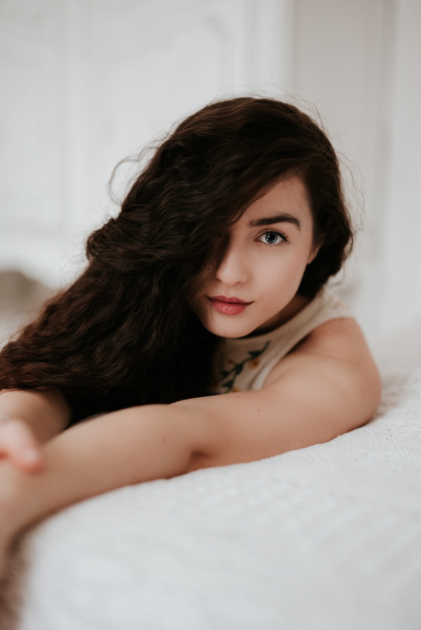 Portret młodej kobiety z długimi, ciemnymi włosami zasłaniającymi część twarzy, leżącej na białej pościeli i patrzącej w obiektyw, ubrana w bluzkę z delikatnym kwiatowym wzorem.
