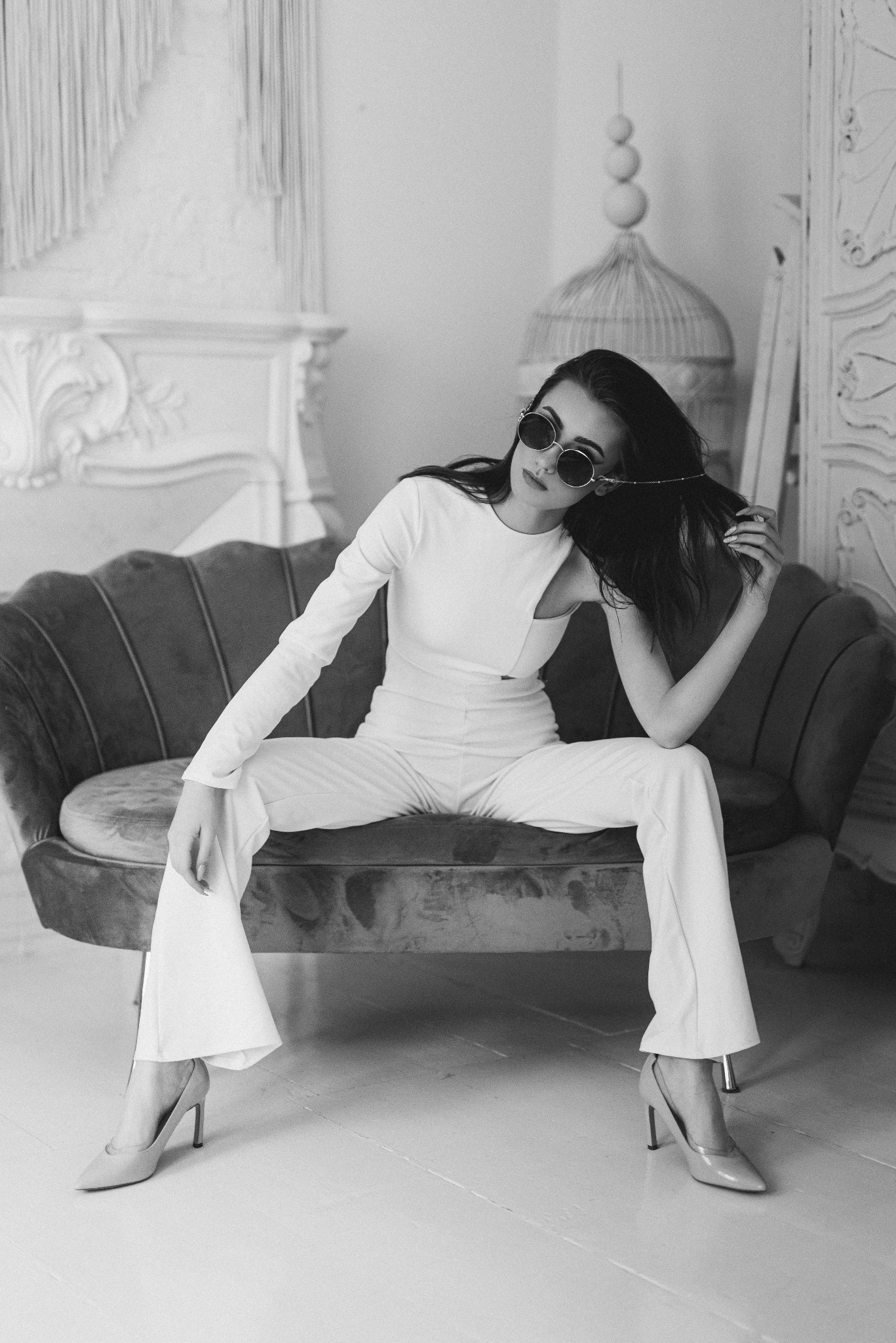 Portret kobiety w białym kombinezonie i okularach przeciwsłonecznych, siedzącej na welurowej sofie w Karlino, w tle dekoracyjny kominek i klatka dla ptaków.