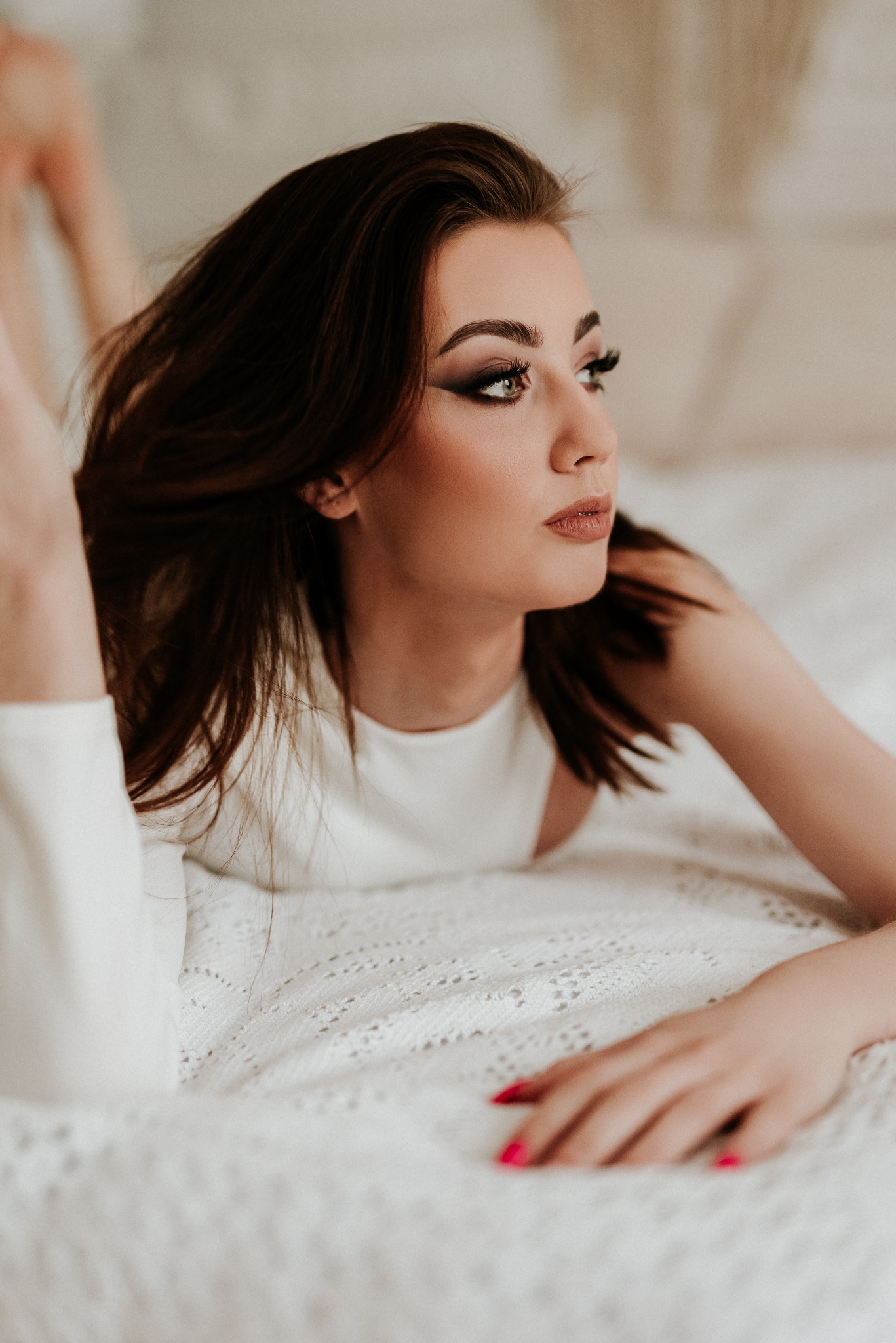 Portret kobiety leżącej na białej, teksturowanej narzucie, z wyraźnym makijażem oczu i różowymi paznokciami, skierowanej wzrokiem w prawo.
