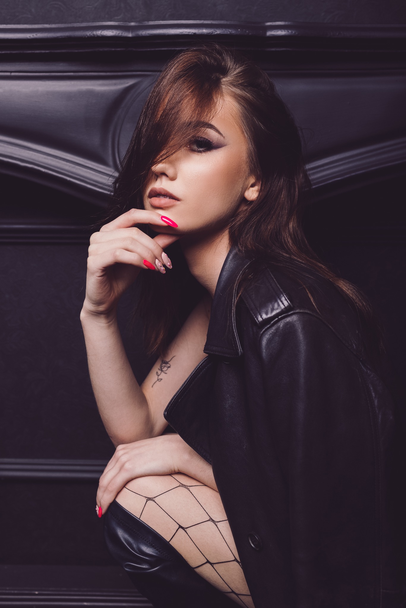 Portret kobiety w czarnej skórzanej kurtce i kabaretkach, z dłonią przy twarzy, ukazujący tatuaż na ramieniu i manicure w odcieniach różu i beżu na tle ciemnej ściany.