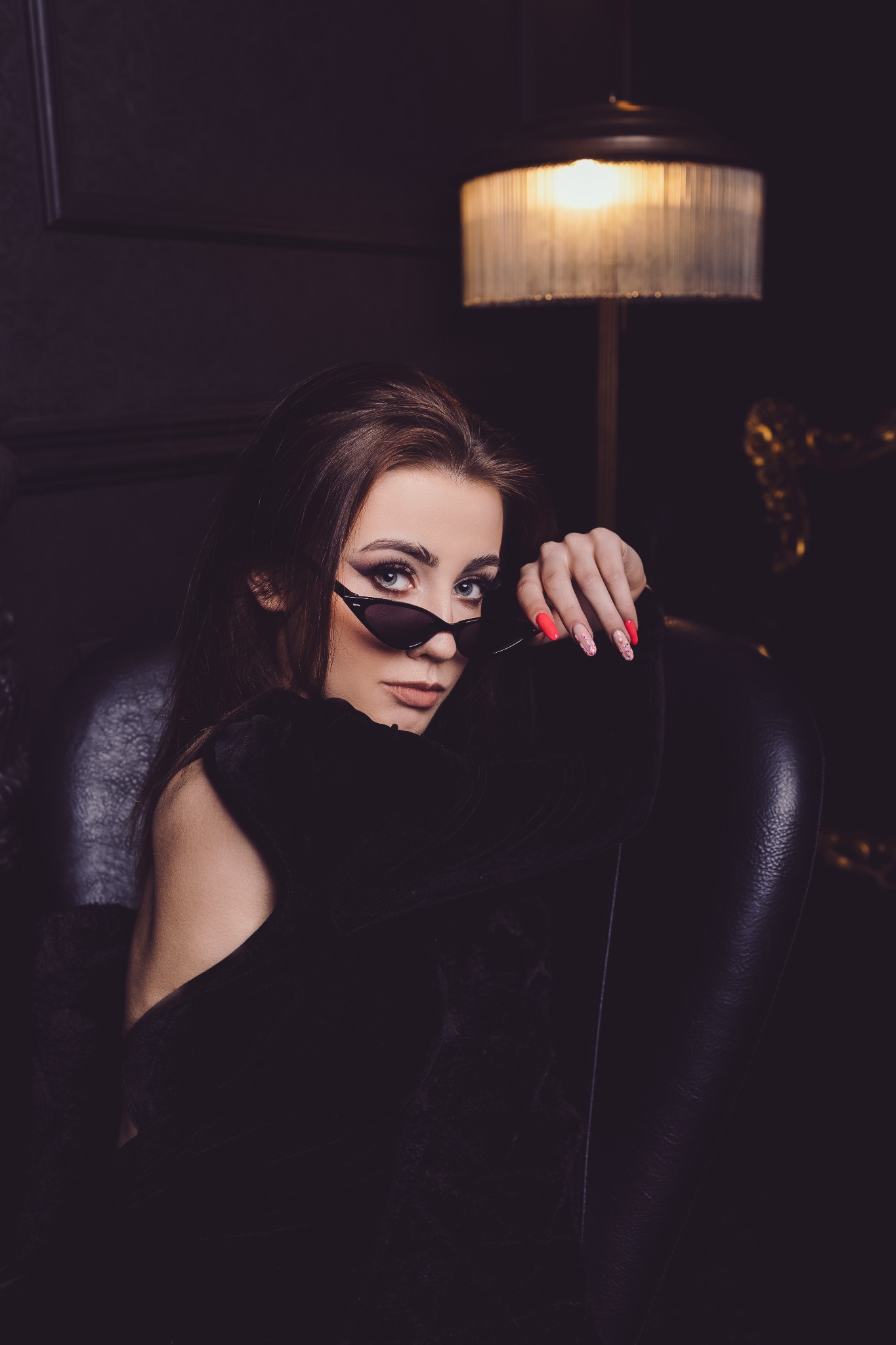 Portret kobiety w czarnym welurowym stroju i okularach w stylu lat 90., siedzącej w skórzanym fotelu na tle ciemnej ściany i lampy.
