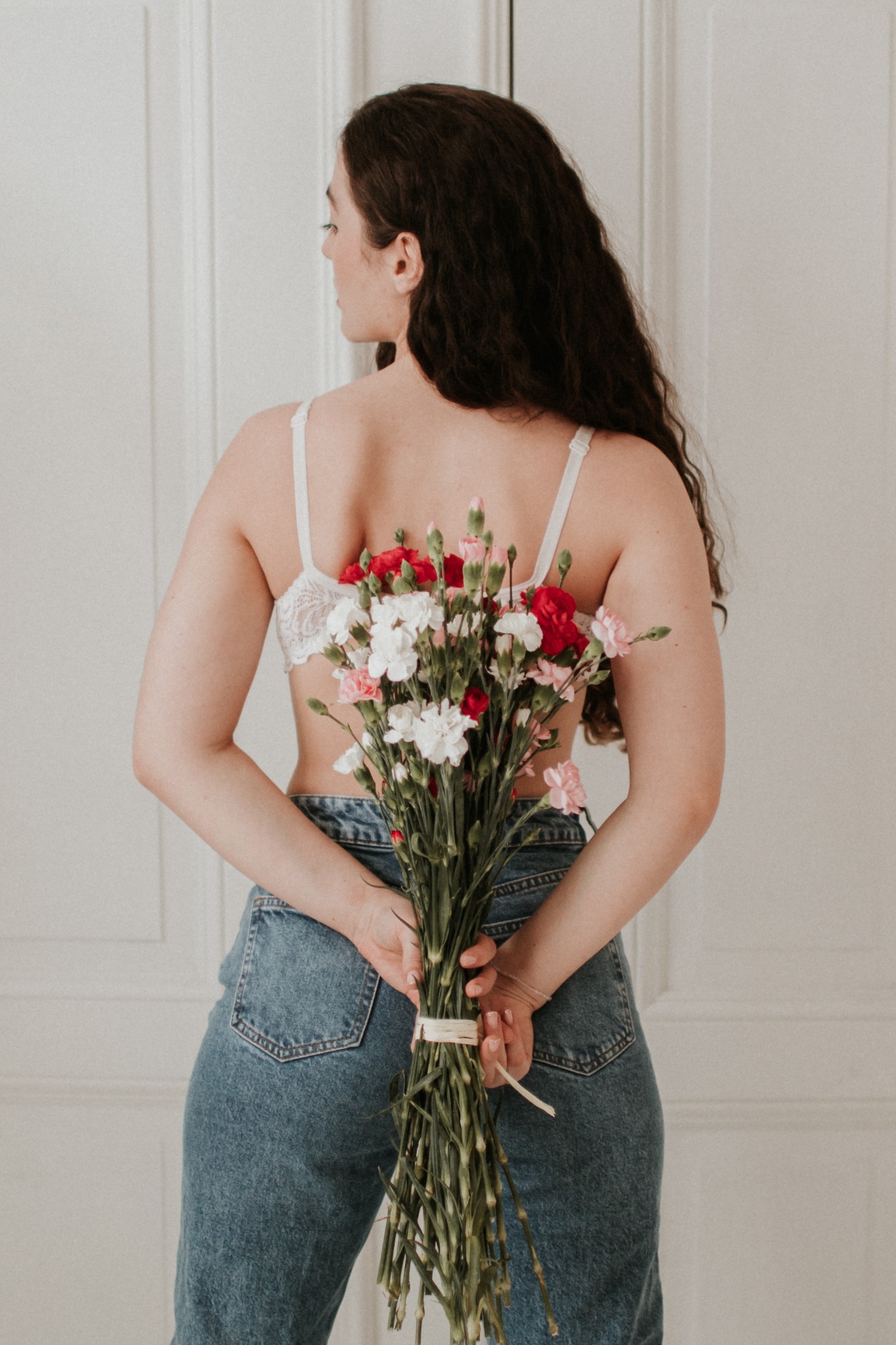 Kobieta odwrócona tyłem, trzymająca za plecami bukiet czerwonych, białych i różowych goździków, ubrana w dżinsy i koronkowy biustonosz na tle białych drzwi.