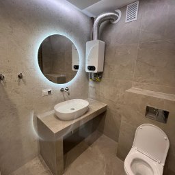 INSTAL BUD Paweł Kalinowski - Beżowa łazienka z dużymi płytkami, okrągłym lustrem LED, owalną umywalką nablatową, podwieszanym WC i gazowym podgrzewaczem wody z elastycznym przewodem.