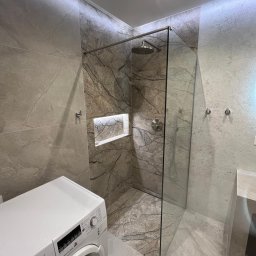 INSTAL BUD Paweł Kalinowski - Nowoczesna łazienka z kabiną prysznicową walk-in, wyłożoną ciemnymi płytkami imitującymi marmur z podświetlaną wnęką. Obok biała pralka Bosch. Jasne, wielkoformatowe płytki na ścianach i podłodze.