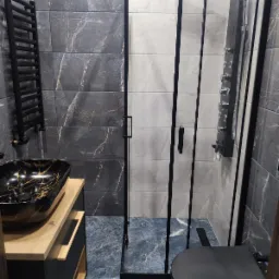 Nowoczesna łazienka z czarną armaturą, marmurowymi płytkami ściennymi i podłogą w odcieniach szarości i błękitu; kabina prysznicowa z czarnymi ramami, umywalka nablatowa w czarnym marmurze ze...