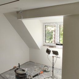 Jackowicz Tomasz - Wnętrze po gładziowaniu w Dębem Wielkim: białe ściany pod skosem, okno, lampa budowlana i odkurzacz. Przygotowanie do malowania.