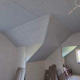 Jackowicz Tomasz - Poddasze z zamontowanymi płytami gipsowo-kartonowymi, widoczne łączenia i wkręty. W tle okno i przejście do innego pomieszczenia. Prace wykończeniowe w surowym stanie.