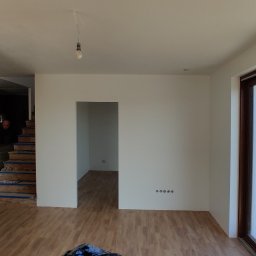 Jackowicz Tomasz - Jasne wnętrze w trakcie prac wykończeniowych: pomalowane ściany, fragment ceglanej ściany z taśmą malarską, widoczne gniazdka elektryczne, drewniane panele podłogowe.