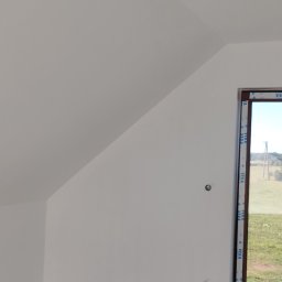 Jackowicz Tomasz - Poddasze po gładziowaniu i malowaniu na biało, widoczne okno z brązową ramą i wystające kable. Szpachlowana podłoga. Ujęcie z dołu, widok na skos dachu.