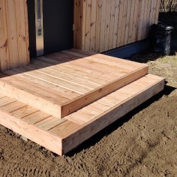 Jackowicz Tomasz - Nowe, drewniane schody wejściowe do domu z boazerią, prowadzące do ciemnych drzwi. Ułożone na ziemi, w otoczeniu trawy i śmieci.