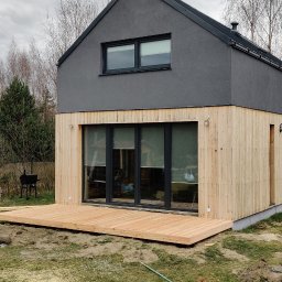 Jackowicz Tomasz - Nowoczesny dom z drewnianą elewacją i tarasem w surowym stanie, otoczony zielenią. Widoczne elementy wykończeniowe i detale architektoniczne.