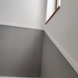 Jackowicz Tomasz - Ściana klatki schodowej po malowaniu: dolna część szara, górna biała, oddzielona listwą. Widoczne fragmenty schodów i okna.