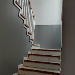 Jackowicz Tomasz - Klatka schodowa w trakcie prac wykończeniowych: pomalowane ściany na szaro, zabezpieczone schody i biała balustrada. Widok z dołu, z bliska.