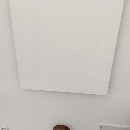 Jackowicz Tomasz - Biały sufit podwieszany z delikatnym oświetleniem punktowym, okrągła lampa sufitowa z drewnianą obudową i klapa rewizyjna w tle, widok z dołu.