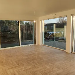 Wykończony pokój z parkietem ułożonym we wzór jodełki francuskiej, widok na zewnątrz przez duże okna przesuwne.