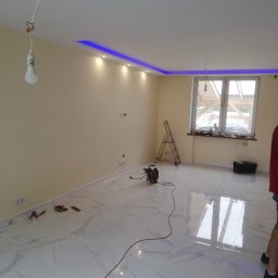 Jackowicz Tomasz - Wnętrze pokoju po remoncie z białymi marmurowymi płytkami podłogowymi, kremowymi ścianami, podwieszanym sufitem z niebieskim oświetleniem LED, widoczne narzędzia i zwisająca żarówka.