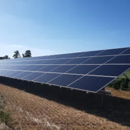 Długa instalacja fotowoltaiczna na polu w Kętrzynie, widok pod kątem, błękitne panele słoneczne i złote ściernisko pod jasnym, błękitnym niebem.