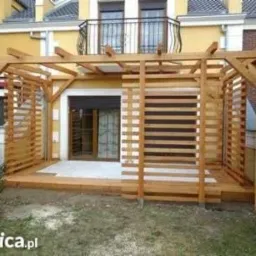 Drewniana konstrukcja z ażurowymi ścianami, przylegająca do budynku z balkonem, tworząca zadaszone patio z drewnianą podłogą.
