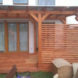 Zadaszony taras z drewnianą podłogą i ażurową ścianką, widoczny fragment budynku z oknami, na tarasie stolik z podstawą maszyny do szycia, w tle nieporządek.