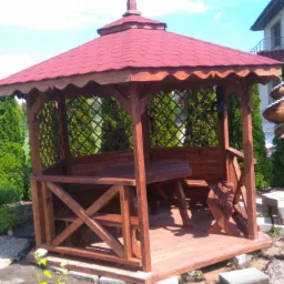 Drewniana altana z czerwonym dachem, wyposażona w stół i ławki, otoczona tujami i elementami dekoracyjnymi ogrodu.