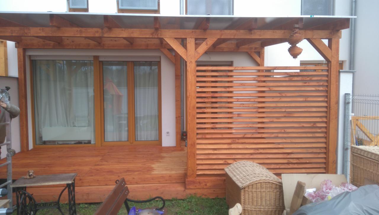 Zadaszony taras z drewnianą podłogą i ażurową ścianką, widoczny fragment budynku z oknami, na tarasie stolik z podstawą maszyny do szycia, w tle nieporządek.