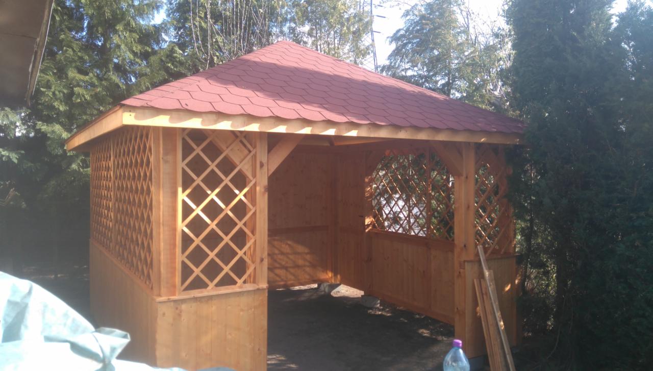 Drewniana konstrukcja altany z czerwonym dachem krytym gontem bitumicznym, częściowo osłonięta kratownicą, na tle zieleni drzew.