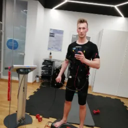 Mężczyzna w kombinezonie do elektrostymulacji EMS, robiący selfie w studiu w Koszalinie. Widoczne elektrody na ciele i sprzęt do ćwiczeń w tle.