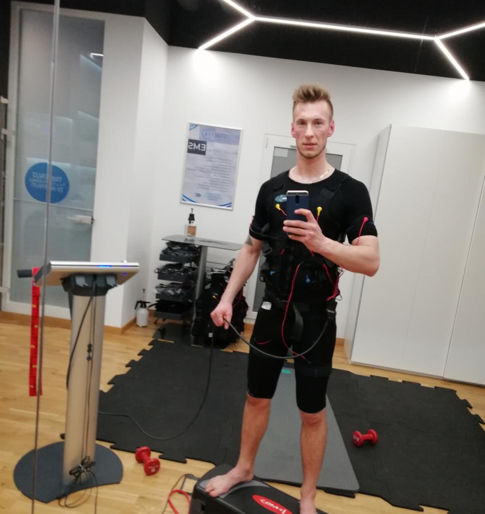 Mężczyzna w kombinezonie do elektrostymulacji EMS, robiący selfie w studiu w Koszalinie. Widoczne elektrody na ciele i sprzęt do ćwiczeń w tle.